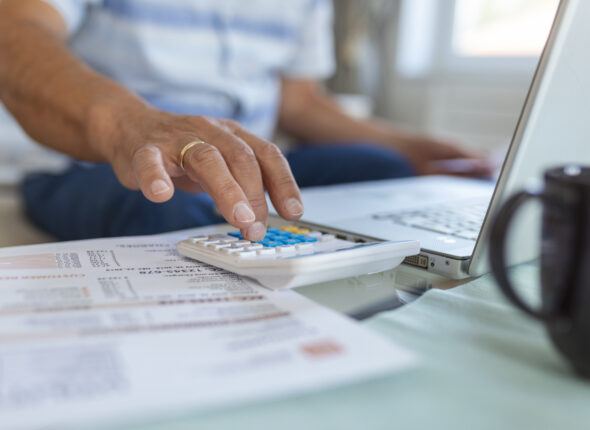 Serious elder senior grey-haired grandpa using calculator counting bank loan payment holding papers at home, focused mature old man doing paperwork calculating bills managing finances sit on sofa